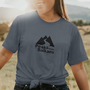 Puede incluir: Una mujer lleva una camiseta gris jaspeada con un gráfico negro de montañas y un camino sinuoso. El texto "Peaks of the Balkans" está impreso debajo del gráfico.