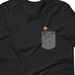 May include: A dark heather grey t-shirt with a pocket featuring a cartoon illustration of a person with a beard and glasses holding a plant.
