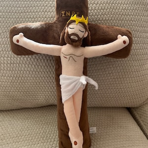 May include: A plush toy depicting Jesus Christ on the cross. The cross is brown and the figure is beige with a white cloth around the waist. The figure has a crown of thorns and a beard. The cross has the inscription "INRI" on the top.