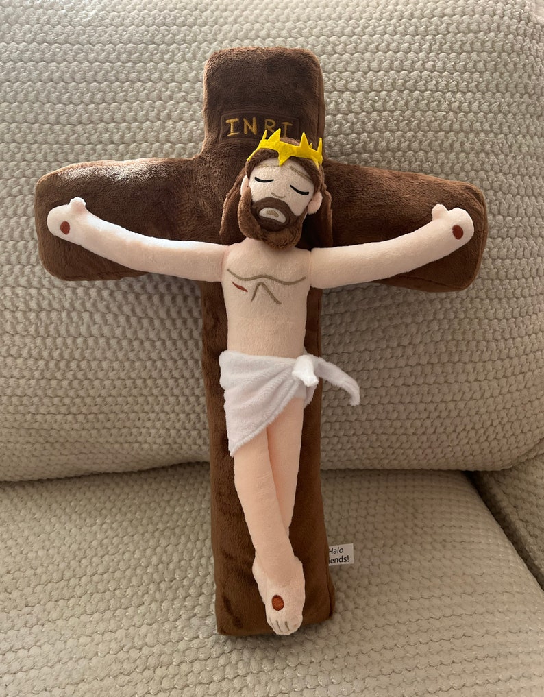 May include: A plush toy depicting Jesus Christ on the cross. The cross is brown and the figure is beige with a white cloth around the waist. The figure has a crown of thorns and a beard. The cross has the inscription "INRI" on the top.