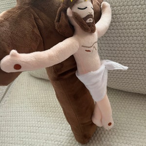 May include: A plush toy depicting Jesus Christ on a brown cross. The toy has a white cloth wrapped around its waist and a yellow crown of thorns on its head. The cross has the letters "INRI" embroidered on it.