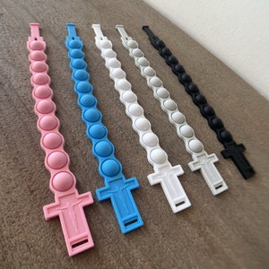 May include: Six silicone bracelets with a cross charm. The bracelets are in pink, blue, white, gray, black, and a darker gray. Each bracelet has a row of 10 round, bubble-like buttons.