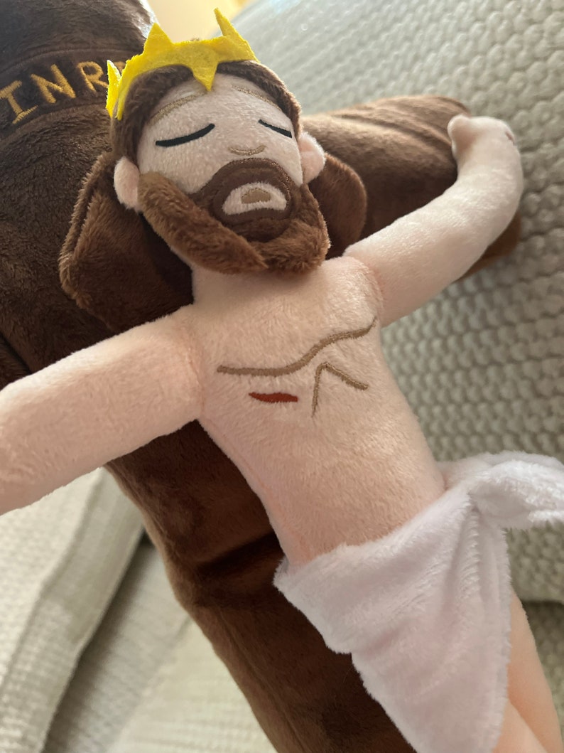 May include: A plush toy depicting Jesus Christ on the cross. The toy is brown and white with a yellow crown and a red wound on the chest. The toy is made of soft, cuddly fabric.