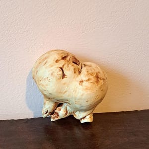 Könnte beinhalten: Eine weiße Keramikskulptur einer stilisierten menschlichen Figur mit einem runden Körper und einer strukturierten Oberfläche. Die Skulptur hat eine braune Glasur und scheint handgefertigt zu sein.