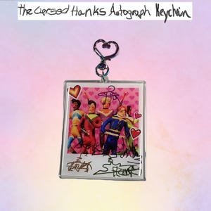 May include: A clear acrylic keychain with a heart-shaped clasp. The keychain features a photo of a group of people in colorful costumes, with signatures at the bottom. The text "the Cersed Hanks Autograph Keychain" is at the top.