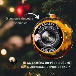Może przedstawiać: Złota bombka choinkowa z motywem obiektywu aparatu i napisem "LA CAMERA DU PERE NOEL". Ozdoba ma grafikę Świętego Mikołaja i napis "IL TE SURVEILLE DEPUIS LE SAPIN!" na rozmytym tle.