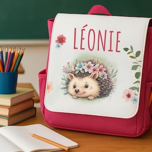 Op de afbeelding: Een magenta rugzak met een witte flap met een illustratie van een egel met een bloemenkrans. De naam "LÉONIE" staat boven de egel. Een potlood zit in de zijzak. Potloden en boeken staan op de achtergrond.