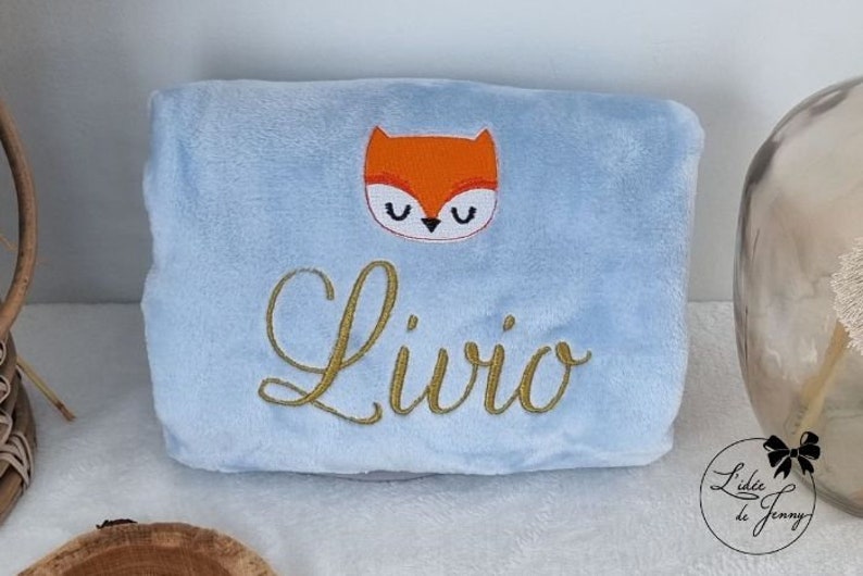 May include: A light blue plush blanket with a gold embroidered name "Livio" and a red fox face with black eyes.