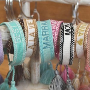 Może przedstawiać: Kolekcja kolorowych, tkanych bransoletek z frędzlami. Bransoletki mają wyszyte różne słowa, w tym "Vibes", "Best", "Ala Vie", "Marra", "Summer", "Mom", "Amina".