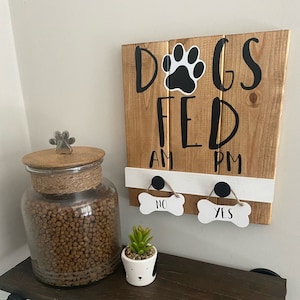 May include: A wooden sign with the text "Dogs Fed" and a paw print graphic. The sign has two toggles that say "AM" and "PM". Below the sign are two bone-shaped toggles that say "No" and "Yes". A glass jar filled with dog food and a small potted plant are on a wooden shelf.