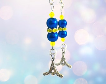 May include: A pair of dangle earrings featuring blue beads, yellow accents, and silver hockey stick charms. The earrings have a hook closure and are designed with a sports theme. The blue beads are a deep, glossy color.