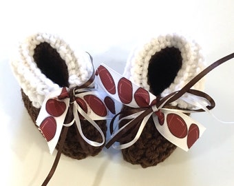 Puede incluir: Un par de botitas de bebé de crochet marrón con ribete de crochet blanco. Las botitas están atadas con una cinta marrón y una cinta blanca con un patrón de fútbol marrón y blanco.