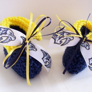 May include: A pair of blue and yellow crocheted baby booties with white ribbon bows. The ribbon bows have a printed design of a blue and yellow mascot.