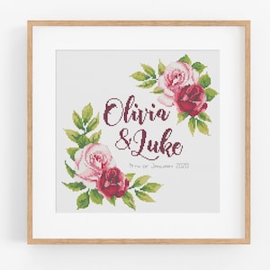 Könnte beinhalten: Ein Kreuzstichmuster mit einem Blumenkranz mit rosa und burgunderfarbenen Rosen. Der Text "Olivia & Luke 11th of January 2020" ist in der Mitte des Kranzes platziert.