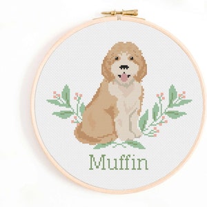 May include: Cross stitch pattern of a brown and white dog with a pink tongue sticking out, sitting in front of green leaves with pink flowers. The dog's name is "Muffin" in green text.