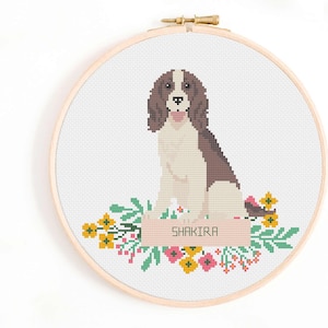 May include: Cross stitch pattern of a brown and white Springer Spaniel dog sitting with a floral wreath around it. The dog's name, "Shakira", is written on a banner.