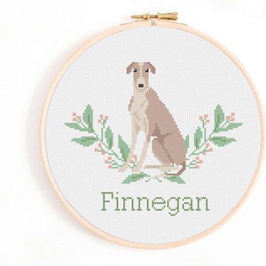 Könnte beinhalten: Kreuzstichmuster eines braunen und weißen Hundes, der mit grünen, belaubten Zweigen und rosa Beeren auf beiden Seiten sitzt. Der Name "Finnegan" ist unter dem Hund gestickt.
