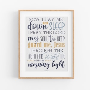 Può includere: Una stampa incorniciata con una cornice in legno chiaro. La stampa presenta il testo "NOW I LAY ME DOWN TO SLEEP" e altri testi religiosi in un mix di caratteri blu, oro e neri. La stampa è un oggetto decorativo.