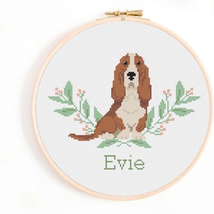 May include: Cross stitch pattern of a brown and white basset hound dog with green leaves and pink flowers. The dog is sitting and the name "Evie" is written below the dog.