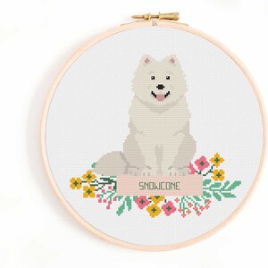 May include: Cross stitch pattern of a white Samoyed dog with a pink nose and tongue, sitting on a pink and green floral banner with the name "SNOWCONE" written on it.