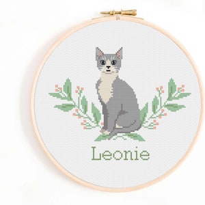 May include: Cross stitch pattern featuring a grey and white cat with green leaves and pink flowers. The name Leonie is stitched below the cat.