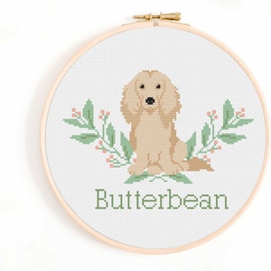 May include: Cross stitch pattern of a golden retriever dog with green leaves and pink berries. The dog is sitting and the text "Butterbean" is below the dog.