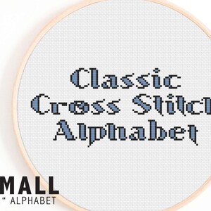 May include: A close-up of a white embroidery hoop with a blue and black cross stitch design that reads "Classic Cross Stitch Alphabet". The hoop is labeled "SMALL <1" ALPHABET".
