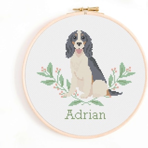 May include: Cross stitch pattern of a dog with black, white, and brown fur sitting with green leafy branches and pink berries on either side. The name "Adrian" is stitched below the dog.