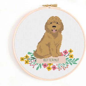 Könnte beinhalten: Ein Kreuzstich-Stickrahmen mit einem goldbraunen Hund mit dem Wort "BUTTERNUT" auf einem Banner. Der Hund sitzt über einem Blumenarrangement mit gelben, rosa und grünen Akzenten. Der Rahmen ist aus Holz.