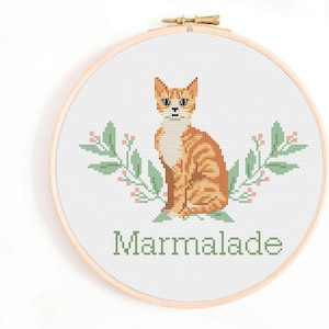 May include: Cross stitch pattern featuring an orange tabby cat with green leaves and pink flowers. The text "Marmalade" is below the cat.