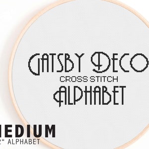 May include: A white embroidery hoop with a black and white cross stitch pattern that reads "Gatsby Deco Cross Stitch Alphabet". The text is in a bold, art deco font. The hoop is labeled "Medium 1-2" Alphabet".