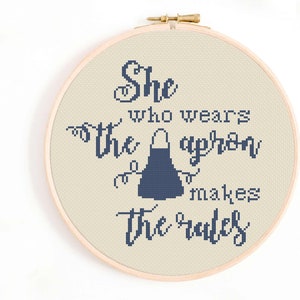 May include: A white embroidery hoop with a beige fabric background. The fabric has a blue embroidered image of an apron and the text "She who wears the apron makes the rules".