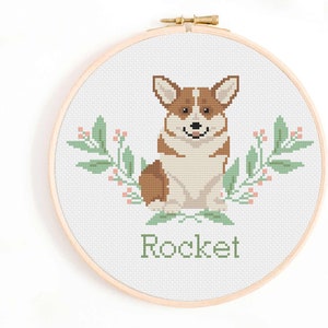 Può includere: Schema di punto croce di un cane corgi marrone e bianco con foglie verdi e fiori rosa. Il nome "Rocket" è ricamato sotto il cane.