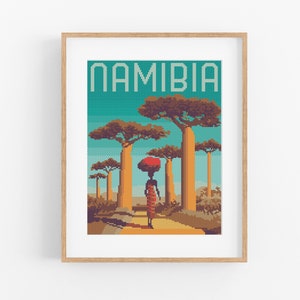 May include: A framed cross-stitch design featuring a woman walking through a forest of tall, orange trees. The trees are surrounded by a blue sky and the text "NAMIBIA" is at the top of the design.