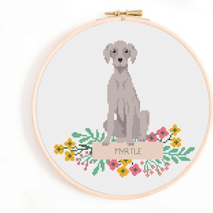 May include: A cross-stitch embroidery hoop featuring a gray dog sitting above a floral arrangement. The dog's name, "MYRTLE," is stitched on a banner. The flowers are pink, yellow, and green, set against a white background.