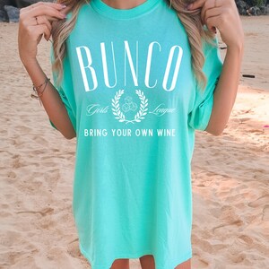May include: A turquoise blue t-shirt with the text "Bunco Girls League" and "Bring Your Own Wine" in white.