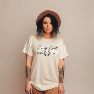 May include: A white t-shirt with the text "Dicey Girls Bunco Club" in black lettering with a wreath design. The model is wearing a brown hat and has tattoos on her arms.