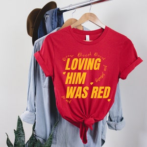 May include: Red t-shirt with the text "Loving Him Was Red" in yellow lettering. The text is surrounded by the word "Record" written multiple times in a smaller font.