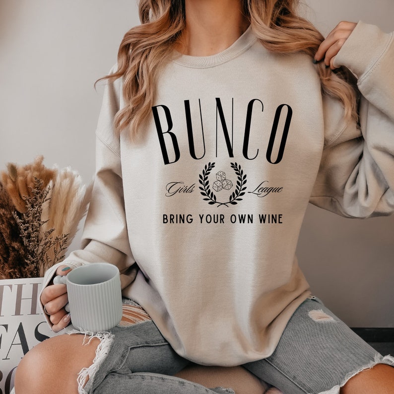 May include: A beige sweatshirt with the text "BUNCO Girls League Bring Your Own Wine" printed on the front. The text is in black and features a wreath with dice in the center.