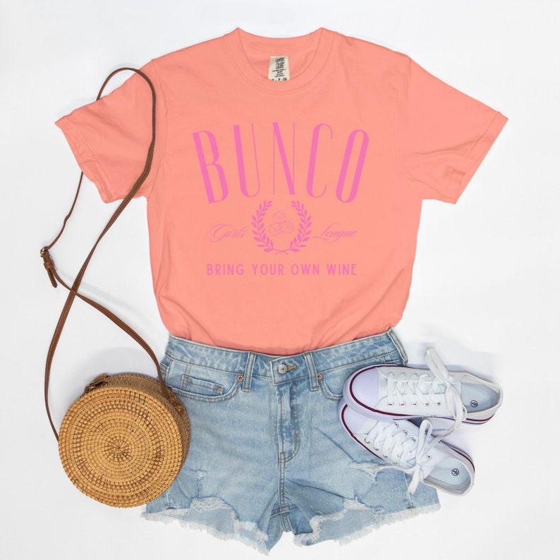 May include: A peach colored t-shirt with the text "Bunco Girls League" and "Bring Your Own Wine" in pink lettering.