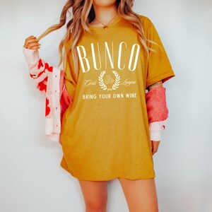 May include: A mustard yellow t-shirt with the text "Bunco Girls League Bring Your Own Wine" in white. The text is surrounded by a wreath with flowers.