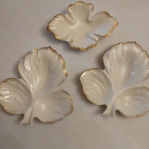May include: Three white leaf-shaped dishes with gold edging. The dishes have a glossy finish and are arranged on a light-coloured surface. The leaf design is detailed with visible veins.
