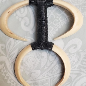 May include: A pendant necklace featuring two curved, off-white tusk-like shapes connected by a central black wrapped section. The necklace cord is black. The design is symmetrical and appears handcrafted.