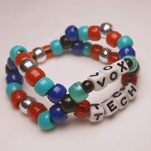 Peut inclure: Deux bracelets de perles. L'un porte le mot "VOX" et l'autre "TECH" en perles blanches. Les bracelets sont faits de perles color&eacute;es rouges, bleues, turquoise, argent&eacute;es et noires.