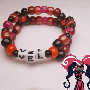 Peut inclure: Un bracelet de perles avec le mot "VEL" &eacute;pel&eacute; en perles de lettres blanches. Le bracelet pr&eacute;sente un m&eacute;lange de perles rouges, noires, oranges et roses. Un personnage de dessin anim&eacute; aux cheveux rouges et noirs se trouve dans le coin inf&eacute;rieur droit.