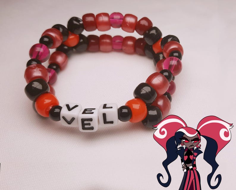 Peut inclure: Un bracelet de perles avec le mot "VEL" &eacute;pel&eacute; en perles de lettres blanches. Le bracelet pr&eacute;sente un m&eacute;lange de perles rouges, noires, oranges et roses. Un personnage de dessin anim&eacute; aux cheveux rouges et noirs se trouve dans le coin inf&eacute;rieur droit.