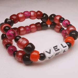 Peut inclure: Un bracelet de perles avec les lettres "VEL" en perles blanches. Le bracelet est compos&eacute; de perles rouges, noires, oranges et roses. Un accessoire fait &agrave; la main.