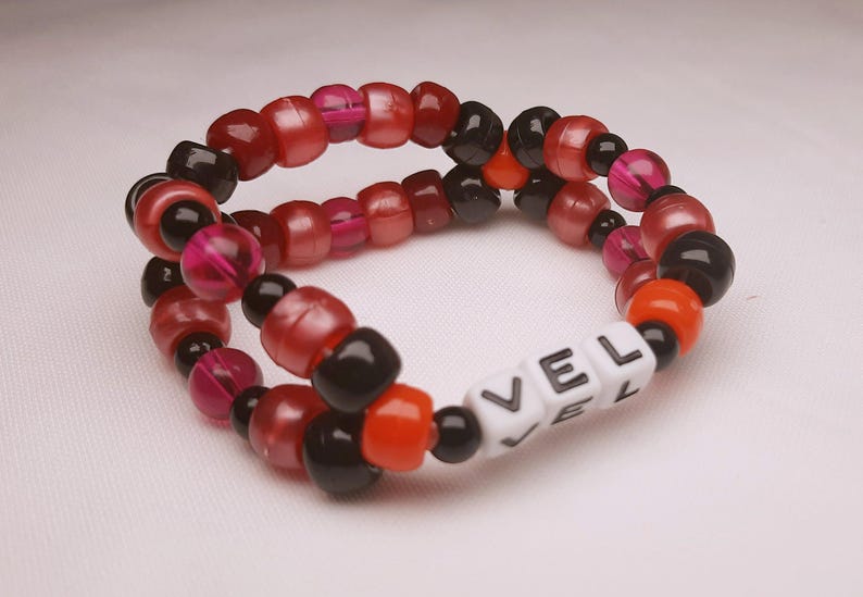 Peut inclure: Un bracelet de perles avec les lettres "VEL" en perles blanches. Le bracelet est compos&eacute; de perles rouges, noires, oranges et roses. Un accessoire fait &agrave; la main.