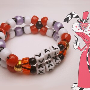 Peut inclure: Un bracelet de perles &agrave; deux rangs avec le mot "VAL XXX" &eacute;crit en perles blanches. Le bracelet est compos&eacute; de perles rouges, oranges, noires, blanches, violettes et dor&eacute;es. Un personnage de dessin anim&eacute; en rouge et noir est en arri&egrave;re-plan.