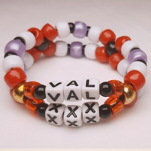 Peut inclure: Deux bracelets de perles. Le bracelet sup&eacute;rieur pr&eacute;sente des perles rouges, blanches et violettes. Le bracelet inf&eacute;rieur comporte des perles de lettres blanches &eacute;pelant "VAL" et "XXX". Les bracelets sont faits de petites perles rondes de diff&eacute;rentes couleurs.